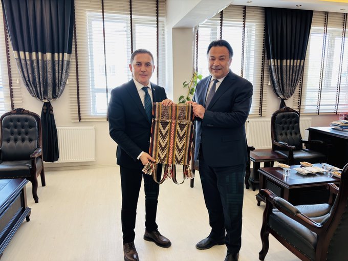 First image shows two men in formal suits one with gray hair wearing a light jacket and the other in dark suit standing in an office holding a large woven patterned bag with geometric designs the office has large windows with blinds a Turkish flag on a pole a portrait on the wall a wooden desk with chairs and a potted plant nearby. Second image depicts two men in dark suits one with short dark hair and the other with similar features standing in a similar office holding a woven patterned bag and a small potted plant the setting includes curtains large windows a wooden desk with chairs and office supplies visible.