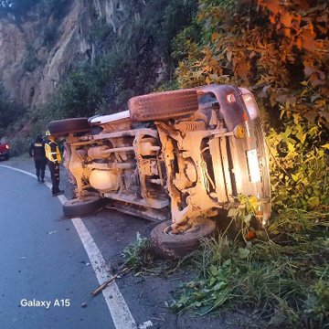 First image displays an overturned red pickup truck on a steep roadside embankment surrounded by lush green vegetation and rocks under dim evening light. The vehicle rests on its side with exposed undercarriage wheels and frame visible amid scattered debris. A firefighter in black uniform and yellow vest stands nearby examining the scene. Second image shows a white ambulance with red stripes parked on the road near the accident site. Several people including women in colorful traditional Mayan huipil blouses and skirts gather around. A firefighter in yellow uniform and helmet directs activities while locals including children observe.