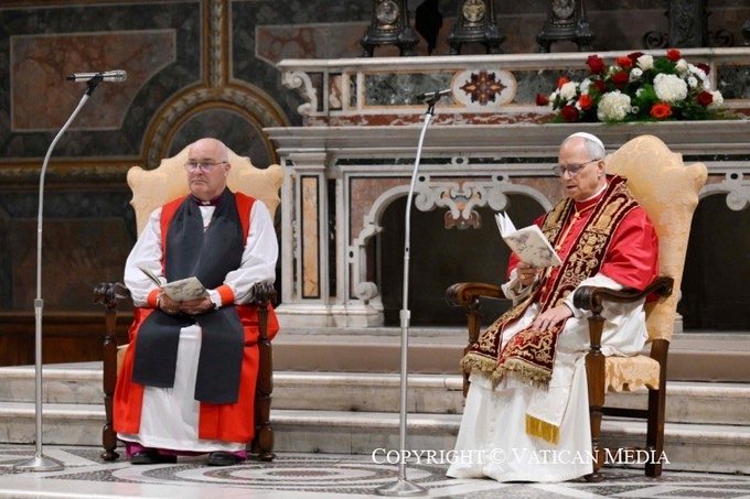 Two elderly men in ornate red and white ecclesiastical vestments sit side by side on cushioned armchairs in a grand chapel with vaulted ceilings, ornate gold decorations, microphones, and floral arrangements. The man on the left holds a book and wears a black cassock under red. The man on the right holds an open book and wears red vestments with gold trim. A copyright notice from Vatican Media appears at the bottom.