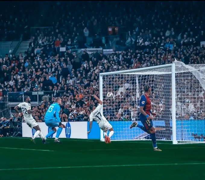 Four soccer players in action near the goal during a match at a stadium with a crowd in the background. A Barcelona player in blue and red jersey jumps to head the ball towards the net while a Real Madrid defender in white jersey attempts to block. The goalkeeper in green jersey dives to save but misses. The scene captures the moment of scoring with the ball entering the goal, surrounded by advertising boards including HP logo.