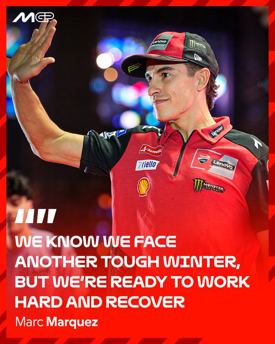 Marc Marquez stands in red and black Ducati racing uniform with Shell Ducati Lenovo logos on chest and arms, wearing a red Red Bull cap, raising his right hand in a fist pump gesture, smiling confidently with focused eyes, against a blurred outdoor nighttime background with bokeh lights and people in distance.