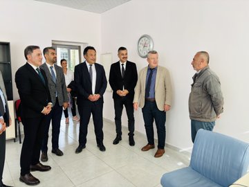 First image shows a group of about 15 professionals in suits and formal attire standing on a paved area outside a white building with green signs reading Tunceli Halk Egitimi Merkezi and Halil Rufai, under a clear sky with trees nearby. Second image depicts six men in suits seated around a black table in an indoor room with beige walls, decorative items like clocks and vases on shelves, water bottles and documents on the table. Third image features eight people including men in suits and a woman in a white top standing in a white-walled room with a clock, blue chairs, and a door. Fourth image shows ten individuals in business attire gathered in a similar indoor space with a clock, red chair, and blue sofa.