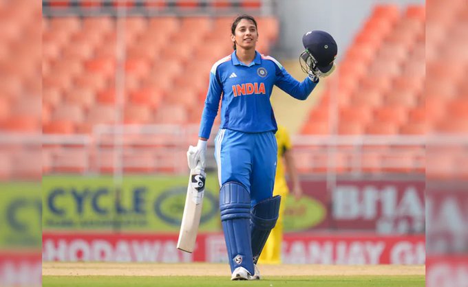 Female cricketer in blue India uniform with white pads and gloves stands on green field holding white bat in right hand and blue helmet in left hand raised arm, stadium seating and sponsor banners in blurred background.