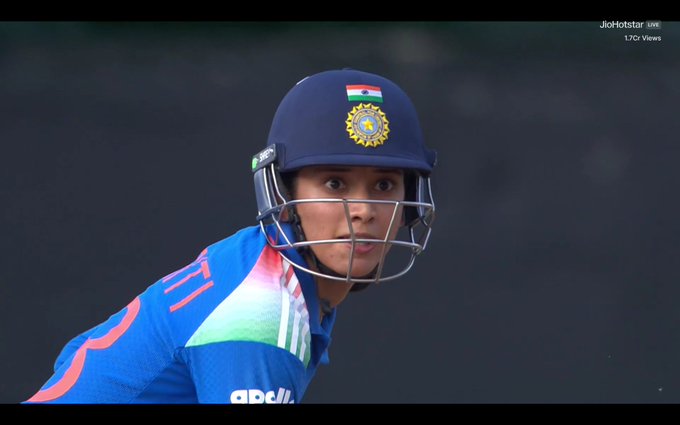 A female cricketer in a side profile view, wearing a blue batting helmet with the Indian tricolor emblem on the front and a protective faceguard, a blue jersey with tricolor stripes on the shoulders and sponsor logos including Adidas, positioned as if ready to bat on a cricket field with a blurred green background.