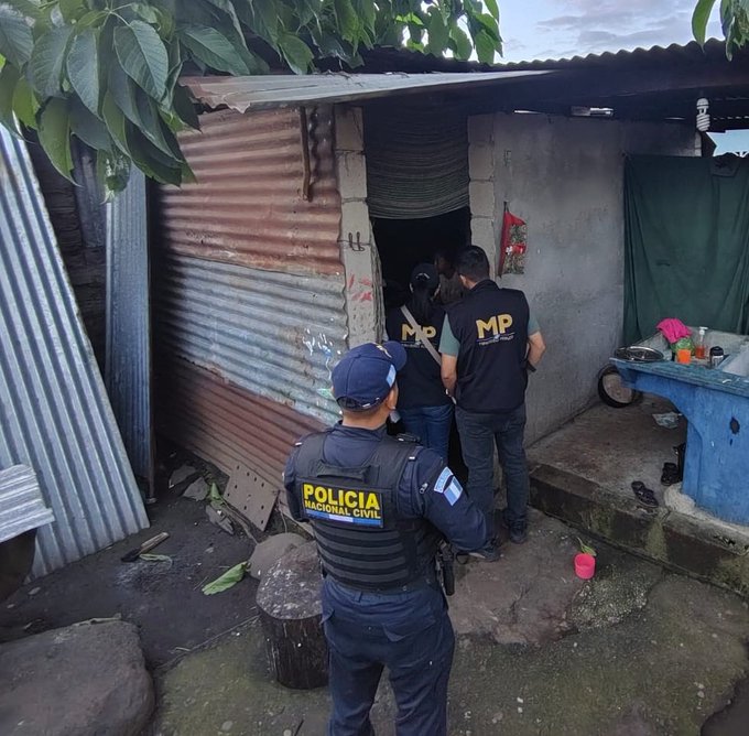 Several police officers in blue uniforms and vests marked POLICIA NACIONAL CIVIL stand outside a rustic structure made of corrugated metal sheets and concrete blocks in a rural setting with greenery and dirt ground. Two officers wearing black shirts with MP on the back escort a man in dark clothing from the doorway. Another officer in a cap and vest stands nearby observing. A blue plastic chair and table with items like a cup are visible in the foreground near the entrance. The scene appears to be an official operation with personnel positioned around the property.