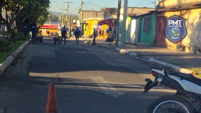 Street view in an urban neighborhood with colorful buildings including yellow and green structures, power lines overhead, trees along the roadside, several police officers in blue uniforms standing and walking, a white motorcycle parked with a traffic cone nearby, workers performing road repairs on the asphalt surface, and the PMT Villa Nueva logo overlaid in the corner.