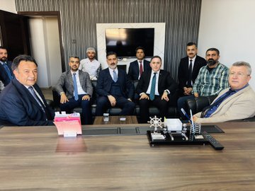 First image shows a group of men in suits and formal attire seated around a conference table in a modern office with a large TV screen on the wall and nautical decor items like a ship wheel model on the table, engaged in a professional meeting. Second image depicts a diverse group of people including chefs in white uniforms and officials in business suits standing around a long table laden with various baked goods such as cakes, pastries, baklava, cookies, and breads in a kitchen setting. Third image captures two individuals, one in a suit and one in chef attire, holding a certificate with a logo while surrounded by staff in uniforms near trays of fried pastries in an industrial kitchen. Fourth image features two men, one in casual pants and shirt and another in a suit, holding a document together with bakery staff in uniforms and headwear standing nearby trays of sweets and pastries on a counter in a kitchen environment.
