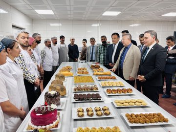 First image shows a group of men in suits and formal attire seated around a conference table in a modern office with a large TV screen on the wall and nautical decor items like a ship wheel model on the table, engaged in a professional meeting. Second image depicts a diverse group of people including chefs in white uniforms and officials in business suits standing around a long table laden with various baked goods such as cakes, pastries, baklava, cookies, and breads in a kitchen setting. Third image captures two individuals, one in a suit and one in chef attire, holding a certificate with a logo while surrounded by staff in uniforms near trays of fried pastries in an industrial kitchen. Fourth image features two men, one in casual pants and shirt and another in a suit, holding a document together with bakery staff in uniforms and headwear standing nearby trays of sweets and pastries on a counter in a kitchen environment.
