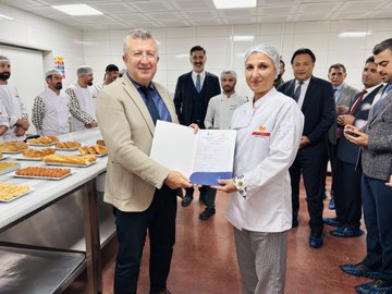 First image shows a group of men in suits and formal attire seated around a conference table in a modern office with a large TV screen on the wall and nautical decor items like a ship wheel model on the table, engaged in a professional meeting. Second image depicts a diverse group of people including chefs in white uniforms and officials in business suits standing around a long table laden with various baked goods such as cakes, pastries, baklava, cookies, and breads in a kitchen setting. Third image captures two individuals, one in a suit and one in chef attire, holding a certificate with a logo while surrounded by staff in uniforms near trays of fried pastries in an industrial kitchen. Fourth image features two men, one in casual pants and shirt and another in a suit, holding a document together with bakery staff in uniforms and headwear standing nearby trays of sweets and pastries on a counter in a kitchen environment.