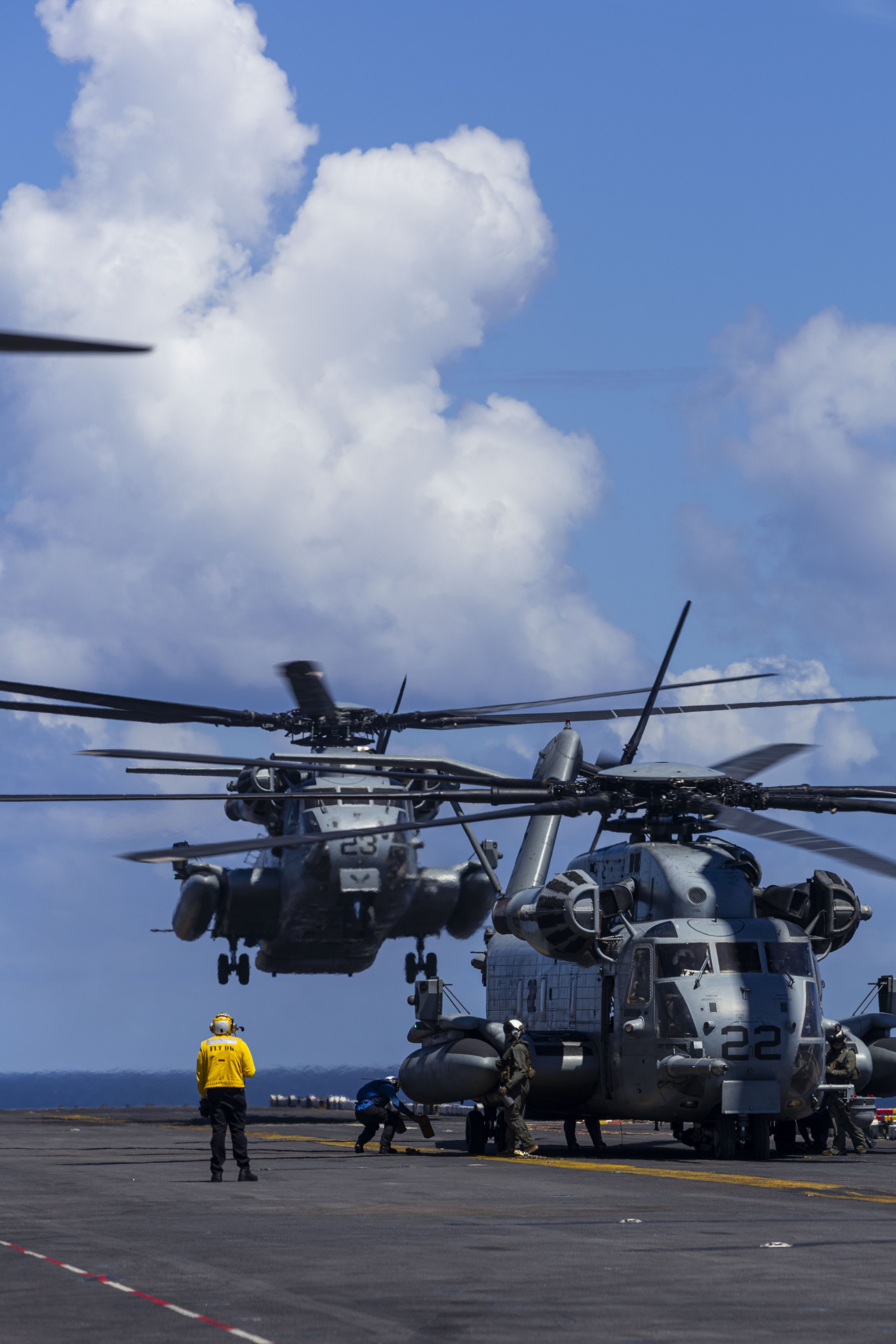 O CH-53E Super Stallion paira sobre um segundo helicóptero no convés de voo de um navio enquanto a tripulação do convés de voo realiza manutenção.