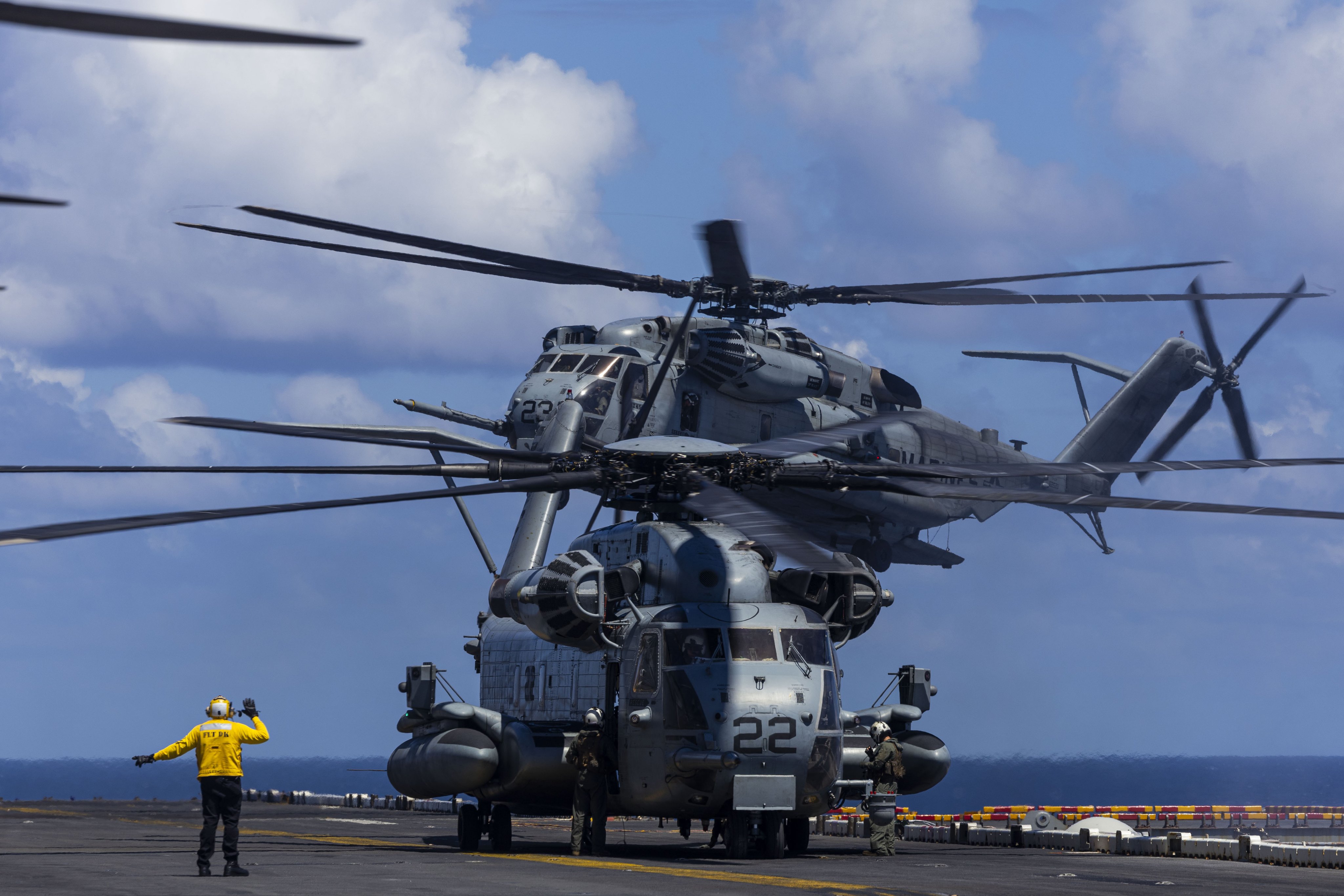 CH-53E Super Stallion lifts off from the flight deck as another helicopter idles below, guided by a Sailor in a yellow jersey.