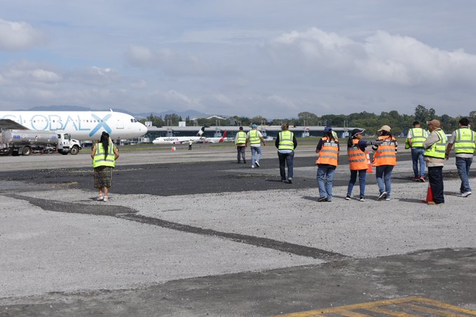 Airport tarmac scene with several workers wearing orange safety vests and helmets walking near a large white airplane with blue and red markings on its side. Additional white aircraft visible in the background parked on the apron. Trucks and equipment positioned nearby under partly cloudy skies with mountains in the distance. Ground crew in various colored clothing including orange and green pants gathered around the area.