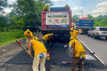 Multiple images show groups of workers in yellow uniforms and helmets performing asphalt patching on a rural road. They use shovels, rakes, and a green compactor machine to spread and level hot asphalt from red dump trucks marked with COVIAL and CAMVIO logos. Surrounding elements include lush green trees, parked vehicles like sedans and SUVs, traffic cones, and a partly cloudy sky. One image focuses on workers loading and spreading material near a truck, another on compacting the surface with a roller, and others capture the team coordinating the repair process.