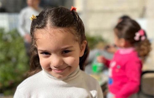 First image shows a young girl with dark hair in pigtails adorned with yellow and red clips smiling while wearing a white top outdoors near green plants with another girl in pink in the background holding a green bottle and an adult figure partially visible. Second image depicts the same girl smiling in a white shirt with plaid pants sitting near a table with bottles and plants in an outdoor setting with another girl in pink nearby. Third image features a young girl with dark hair in a bun wearing a tiara and a pink puffy sleeved dress standing indoors near wooden furniture and decorative walls.