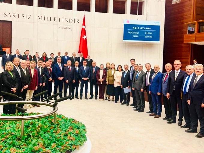 First image shows a large group of about 30 people in formal business attire standing in a semi-circle in a grand assembly hall with white marble walls inscribed with Turkish text meaning Justice is the foundation of the state and The state belongs to the nation, a Turkish flag on the right, a digital display showing AG 2024 in the background, and green plants in the foreground. Second image depicts a smaller group of eight individuals including men and women in suits and dresses posing formally in front of similar marble walls and a Turkish flag, with wooden benches and doors visible in the parliamentary setting.