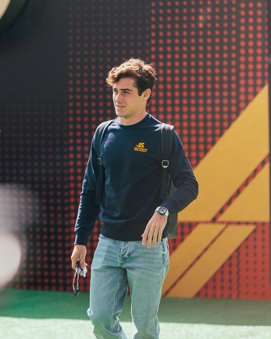 First image: Young man with curly brown hair, smiling slightly, dressed in navy blue hoodie with sponsor logo on chest, black backpack strap over right shoulder, silver watch on left wrist, sunglasses dangling from hoodie neckline, light blue jeans, standing on green grass with hands in pockets, background of large red and yellow abstract panels on a structure. Second image: Man with short reddish hair, wearing blue Alpine racing jacket with white sponsor patches on sleeves and chest, dark pants tucked into white sneakers, standing on green grass in front of a modern entrance framed by black and red panels, with a white car and trash bin visible to the side, green foliage in background.
