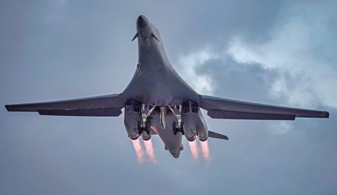 A large military bomber aircraft with swept-back wings and four engines visible under the fuselage takes off from a runway with bright orange flames from the jet exhausts against a cloudy sky. The plane is angled upward showing its underbelly and landing gear partially extended. No people or ground details are visible.