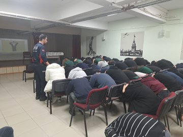 First image shows a person in safety helmet and work clothes holding a red fire extinguisher spraying foam at a small fire in a black container producing smoke on an outdoor paved area surrounded by apartment buildings with a group of people including some in helmets seated and standing watching. Second image depicts a similar outdoor scene with a small flame in a black container on the ground and a person in uniform standing nearby while a large group of students in school uniforms sit on steps around a pavilion area with buildings in background. Third image features a group of teachers and staff including some in red safety vests posing for a photo on steps in front of a gray school building labeled Nuri Vatan Anadolu Lisesi with Turkish flag emblem. Fourth image illustrates an indoor classroom setting with a uniformed instructor standing at front near projector screen addressing rows of seated participants many wearing caps and casual clothes focused on the presentation.