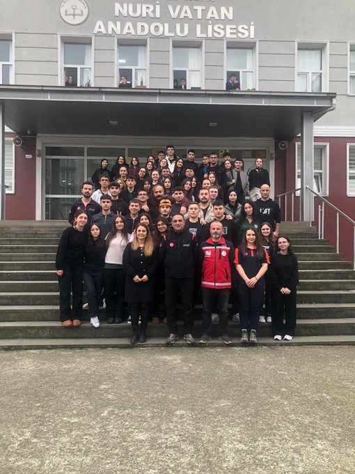 First image shows a person in safety helmet and work clothes holding a red fire extinguisher spraying foam at a small fire in a black container producing smoke on an outdoor paved area surrounded by apartment buildings with a group of people including some in helmets seated and standing watching. Second image depicts a similar outdoor scene with a small flame in a black container on the ground and a person in uniform standing nearby while a large group of students in school uniforms sit on steps around a pavilion area with buildings in background. Third image features a group of teachers and staff including some in red safety vests posing for a photo on steps in front of a gray school building labeled Nuri Vatan Anadolu Lisesi with Turkish flag emblem. Fourth image illustrates an indoor classroom setting with a uniformed instructor standing at front near projector screen addressing rows of seated participants many wearing caps and casual clothes focused on the presentation.