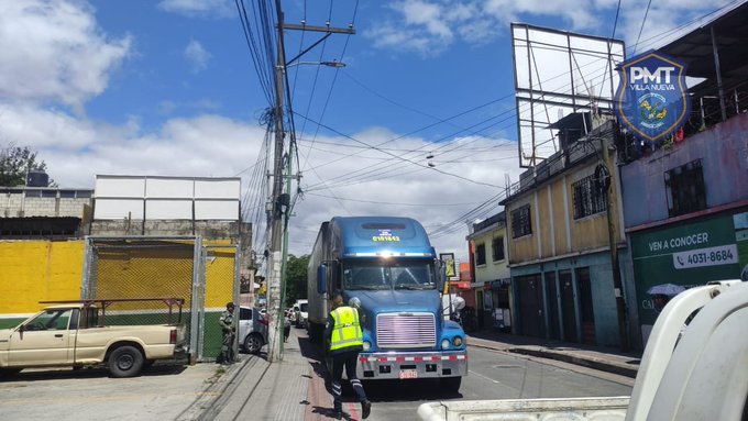 A blue semi-trailer truck with mechanical issues is stopped on a narrow urban street in Villa Nueva, Guatemala, with its hood possibly open for inspection. A traffic officer in a high-visibility yellow vest stands beside the truck, appearing to examine it. Surrounding elements include a beige pickup truck parked nearby, several motorcycles, yellow and green buildings with metal roofs, overhead power lines, a billboard, and a sign for PMT in the background under a partly cloudy sky.
