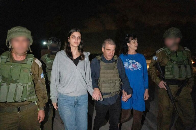 La escena nocturna muestra a cuatro soldados con uniformes militares verdes y chalecos sosteniendo rifles, escoltando a tres individuos: una mujer con sudadera con capucha gris y jeans, un hombre con camisa y chaleco azul, y otra mujer con vestido azul, todos caminando por una carretera con vehículos y luces de fondo, rostros de soldados borrosos.