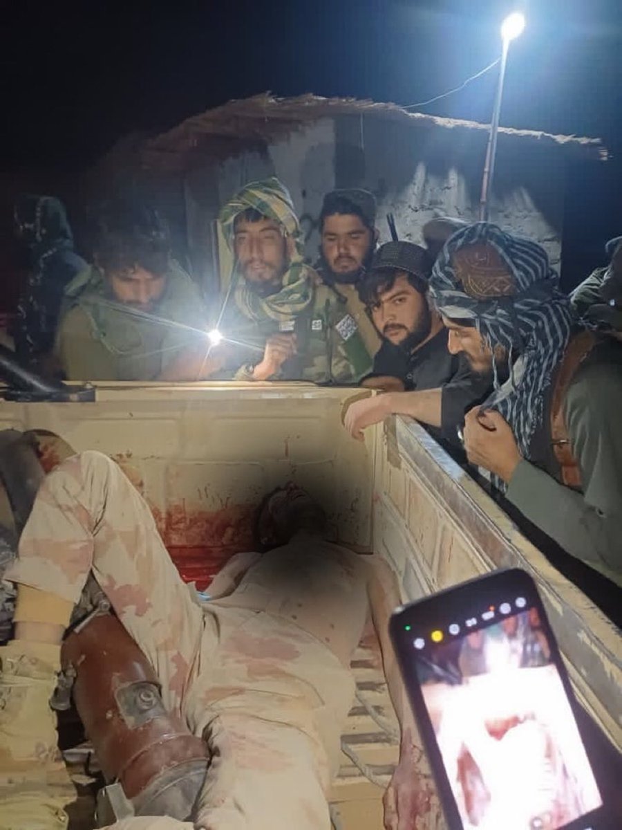 Group of men wearing camouflage uniforms and headscarves, some armed with rifles, gathered around a truck bed at night illuminated by a light. Bloodied bodies in similar uniforms lie on the ground beside the truck, with visible blood stains on clothing and skin. A smartphone screen in the foreground displays a close-up of one injured face. Setting appears to be a damaged outdoor area with rubble.