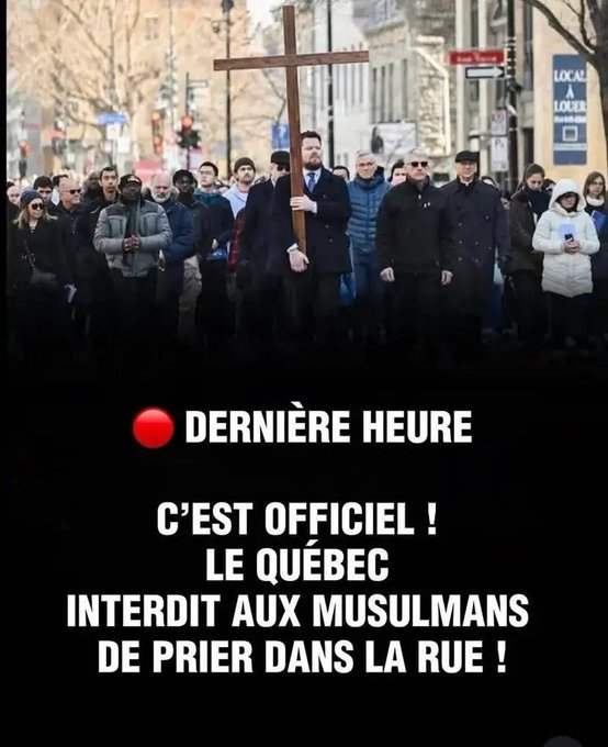 A crowd of diverse individuals stands on a paved urban street lined with bare trees and buildings, holding protest signs and a large wooden cross upright. Participants wear coats, hats, and scarves suitable for cold weather, gathered near a red sign reading Local. The scene appears to be a demonstration with people facing forward in solidarity.