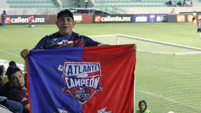 Escena del estadio con campo verde, red de portería y espectadores en el fondo. Hombre con gorra y camiseta sostiene una gran bandera dividida en azul y rojo con el emblema de Atlante Campeón y texto de México. Otras personas visibles cerca del borde del campo. Caliente.mx marca de agua presente en la imagen.