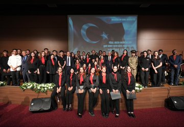 First image shows a man in a dark suit speaking into a microphone at a podium on a stage with a large screen displaying Turkish flags and text Kim Var in Turkish, alongside event title and sponsor logos, wooden paneling, chairs, and musical instruments in the background. Second image depicts several men in suits seated and holding red Turkish flags with white crescent and star emblems in a dimly lit auditorium filled with audience members waving more flags. Third image captures a man in a dark suit shaking hands with a woman in camouflage military uniform and cap, surrounded by young men and women in casual dark clothing and a woman in black attire, near a doorway with beige walls and a guitar leaning against it. Fourth image features a large group of men and women in black suits and red scarves standing on a wooden stage with a blue-lit screen showing Turkish flag and 2023-2024 text, holding certificates, flowers on the floor, and speakers nearby.