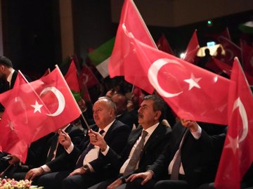First image shows a man in a dark suit speaking into a microphone at a podium on a stage with a large screen displaying Turkish flags and text Kim Var in Turkish, alongside event title and sponsor logos, wooden paneling, chairs, and musical instruments in the background. Second image depicts several men in suits seated and holding red Turkish flags with white crescent and star emblems in a dimly lit auditorium filled with audience members waving more flags. Third image captures a man in a dark suit shaking hands with a woman in camouflage military uniform and cap, surrounded by young men and women in casual dark clothing and a woman in black attire, near a doorway with beige walls and a guitar leaning against it. Fourth image features a large group of men and women in black suits and red scarves standing on a wooden stage with a blue-lit screen showing Turkish flag and 2023-2024 text, holding certificates, flowers on the floor, and speakers nearby.