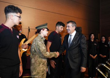 First image shows a man in a dark suit speaking into a microphone at a podium on a stage with a large screen displaying Turkish flags and text Kim Var in Turkish, alongside event title and sponsor logos, wooden paneling, chairs, and musical instruments in the background. Second image depicts several men in suits seated and holding red Turkish flags with white crescent and star emblems in a dimly lit auditorium filled with audience members waving more flags. Third image captures a man in a dark suit shaking hands with a woman in camouflage military uniform and cap, surrounded by young men and women in casual dark clothing and a woman in black attire, near a doorway with beige walls and a guitar leaning against it. Fourth image features a large group of men and women in black suits and red scarves standing on a wooden stage with a blue-lit screen showing Turkish flag and 2023-2024 text, holding certificates, flowers on the floor, and speakers nearby.