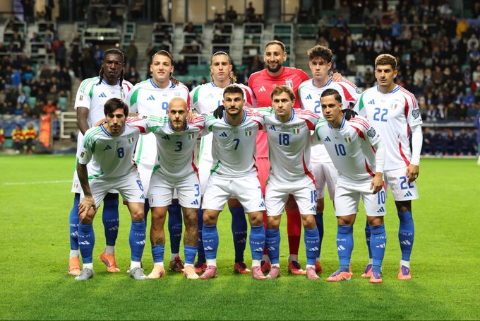 Group of male football players in Italian national team kits with green white and red colors standing in formation on green grass field in stadium with blue seats and crowd visible in background some players kneeling in front others standing behind wearing numbered jerseys and shorts with sponsor logos like Adidas
