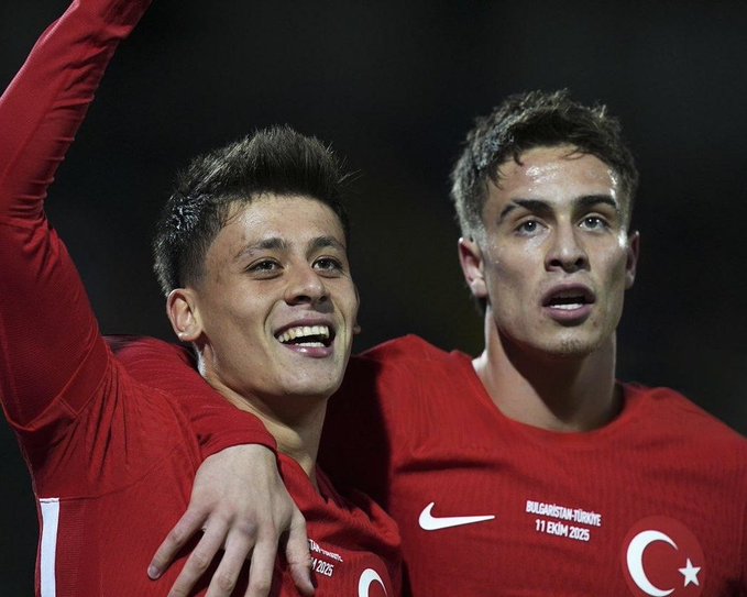 Two young men with short brown hair stand close together on a dark outdoor field, wearing matching red long-sleeve jerseys with white Nike swooshes on the chest, the text 2024-2025 below, and a Turkish flag emblem on the left sleeve, one player raises his right arm upward while gripping the others shoulder with his left hand, both have wide smiles and focused expressions, the second player has his mouth slightly open.