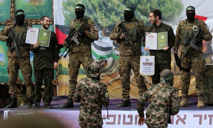 Group of six masked men in green military uniforms and keffiyehs stand on a wooden stage holding rifles, with two in the center displaying green certificates labeled in Arabic, one on the left holding a white bag, backdrop featuring large Palestinian flags, green banners with Arabic text, and a rural landscape setting.