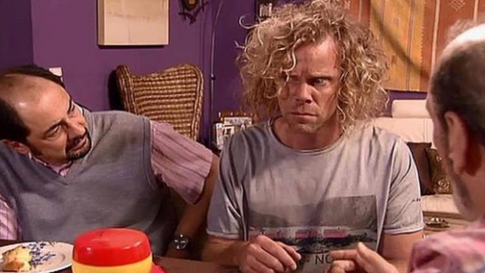 Tres hombres en una sala de estar con paredes moradas y sillas tejidas, uno con cabello rubio rizado y una camiseta gris se ve sorprendido sosteniendo un contenedor rojo, otro hombre calvo con un chaleco gris y una camisa rosa se sienta cerca, un tercer hombre mira hacia otro lado, la escena parece sacada de una comedia de situación con una mesa y elementos decorativos.