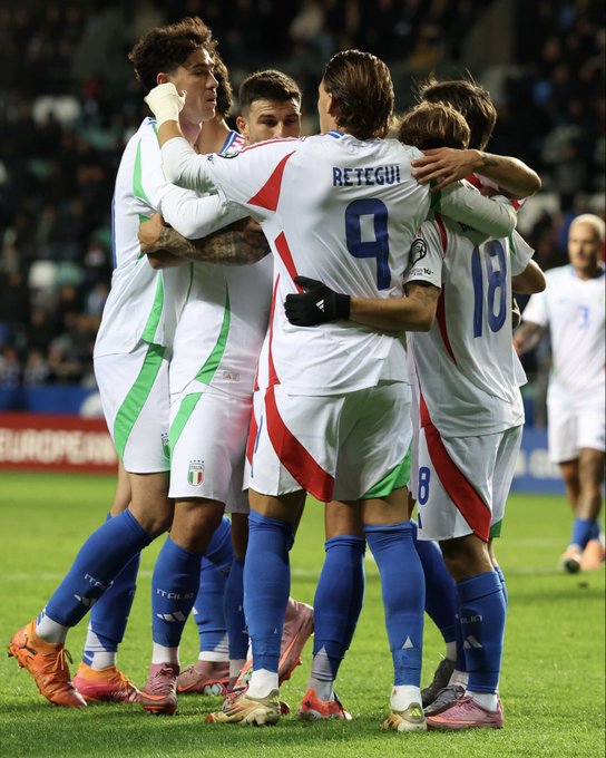 Un grupo de jugadores de fútbol masculinos con uniformes deportivos se apiñan y celebran en un campo de césped verde durante un partido, con un jugador que lleva una camiseta blanca y roja con el número 9 con la etiqueta Retegui, otros con uniformes verdes y blancos con los números 18 y 8, y parches de la bandera italiana visibles en las mangas, los asientos del estadio y las barreras azules en el fondo.