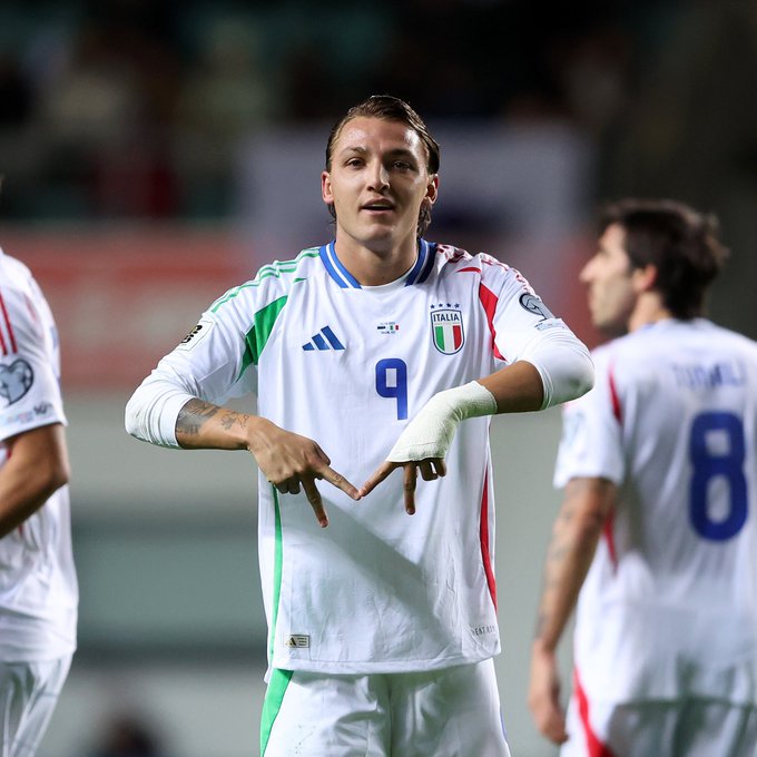 El futbolista Mateo Retegui con la camiseta número 9 de la selección italiana con rayas verdes y rojas y el logotipo de Adidas celebra haciendo un gesto con la mano con ambas manos formando un corazón mientras lleva una muñequera en el brazo derecho y tatuajes visibles en los brazos. Otros dos jugadores italianos con camisetas similares eran 8 y otro se paran cerca en el campo verde con asientos del estadio y pancartas en el fondo bajo las luces del estadio.
