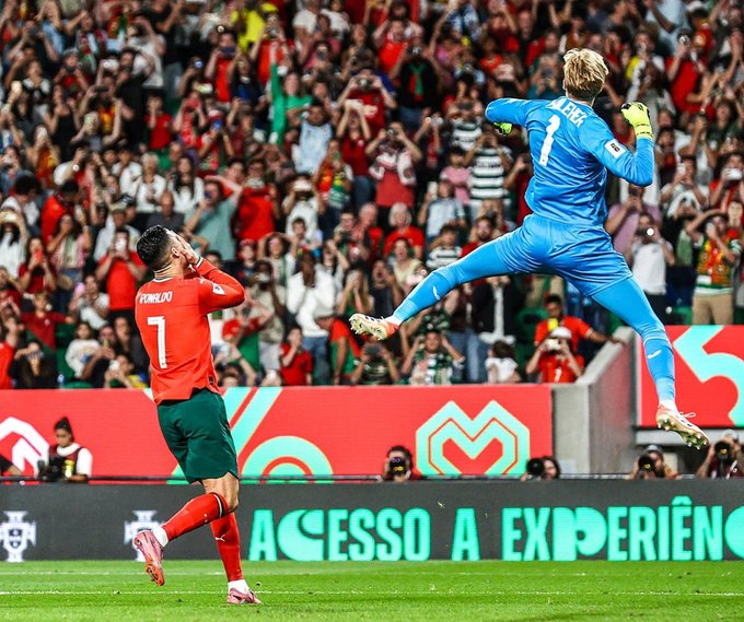 Un portero de fútbol con una camiseta azul con el número 1 salta alto para atrapar un tiro penal con guantes amarillos extendidos, mientras que Cristiano Ronaldo con una camiseta roja de Portugal con el número 7 se para cerca con las manos en la cabeza en señal de decepción, rodeado por un estadio abarrotado con fanáticos vestidos de verde y rojo, y pancartas publicitarias en la parte inferior que dicen Accesos a Experiencia.