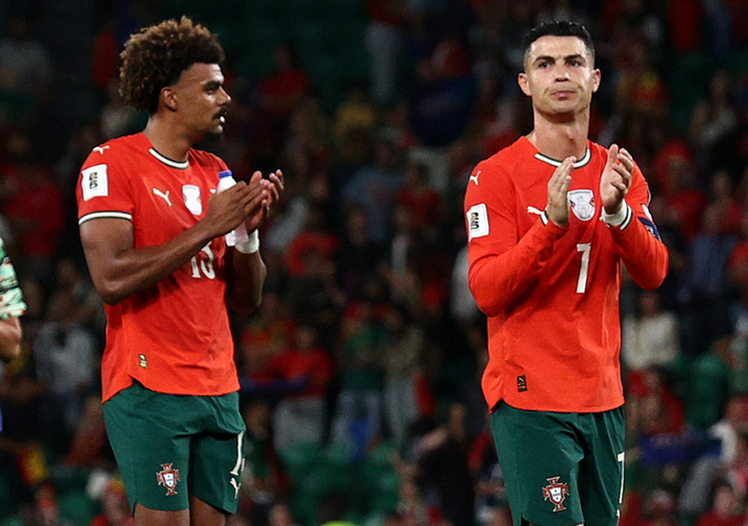 Dos jugadores de fútbol masculinos se paran uno al lado del otro en un campo verde con una multitud borrosa en el estadio en el fondo. El jugador de la izquierda tiene el pelo oscuro rizado, lleva una camiseta roja de manga larga con el número 10, muñequeras negras y pantalones cortos verdes con el parche de la bandera de Portugal. El jugador de la derecha tiene el pelo corto y oscuro, lleva una camiseta roja de manga larga con el número 7, un reloj de pulsera y pantalones cortos verdes con el parche de la bandera de Portugal. Ambos jugadores están aplaudiendo.