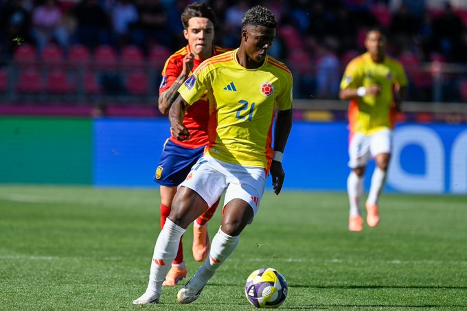Neyser Villareal con la camiseta amarilla y azul de la selección nacional de Colombia número 24 regatea una pelota de fútbol colorida sobre césped verde durante un partido. Un jugador español con uniforme rojo de España con tatuaje en el brazo lo persigue de cerca por detrás. Otros jugadores con uniformes similares visibles en el fondo con asientos de estadio y barreras azules.