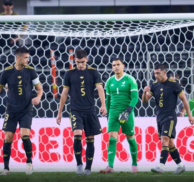 Four soccer players stand near a goal net on a green field with advertising boards behind. Leftmost player in black jersey with number 3 and yellow accents stands with arms at sides. Next player in black jersey with number 5 has hands on hips. Central player is goalkeeper in green jersey with white number 1, arms raised wearing green gloves. Rightmost player in black jersey with number 6 stands relaxed. All wear black shorts and socks with yellow details, white cleats. Net and posts visible, crowd blurred in background.