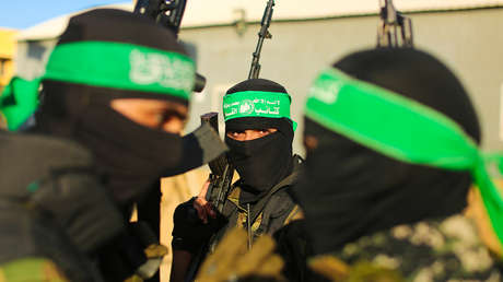 Tres individuos enmascarados que llevan cintas verdes en la cabeza con texto árabe y pasamontañas negros se paran juntos al aire libre contra un edificio beige bajo un cielo despejado. Están vestidos con uniformes de camuflaje de estilo militar y sostienen rifles AK-47. La figura central tiene un brazalete verde y parece enfocada, mientras que las demás parecen alerta.