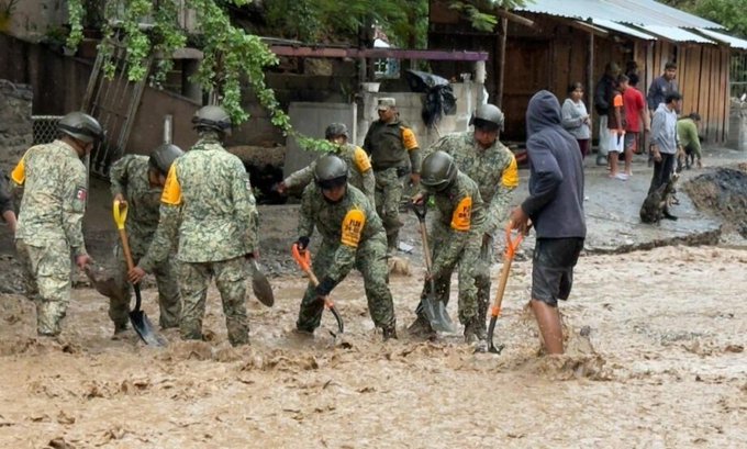 Un grupo de soldados con uniformes de camuflaje con chalecos amarillos y cascos usan palas y herramientas para limpiar el lodo y los escombros de un área inundada cerca de estructuras de madera y árboles, con varios civiles ayudando en el esfuerzo.