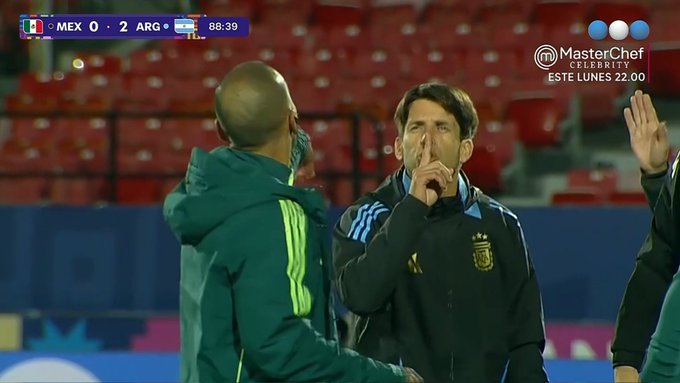 Dos jugadores de fútbol masculinos se paran cerca en un campo de estadio con asientos rojos durante un partido, uno con una chaqueta verde Adidas señalando y hablando animadamente, el otro con una chaqueta azul de Argentina escuchando con la mano levantada. El marcador muestra México 0 Argentina 2 a las 85:39 con banderas, el logotipo de MasterChef y el lunes a las 22:00 horas. El fondo muestra los asientos del estadio y las marcas del campo.