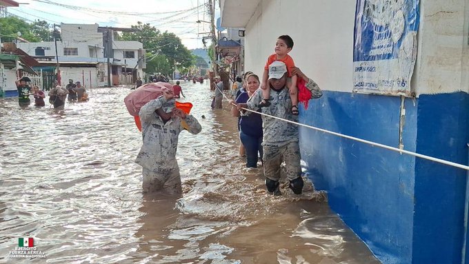 Escena callejera inundada en un pueblo mexicano con agua fangosa que llega a la altura de las rodillas sobre varios adultos y niños que caminan por un edificio de paredes azules con ropa tendida. Un hombre con camisa y gorra azul claro lleva un cubo rojo y una bolsa mientras sostiene la mano de un niño. Otro hombre con camisa gris lleva a un niño sobre sus hombros y sostiene una bolsa roja. Las personas en el fondo llevan bolsas a través del agua cerca de edificios blancos y árboles bajo el cielo nublado.