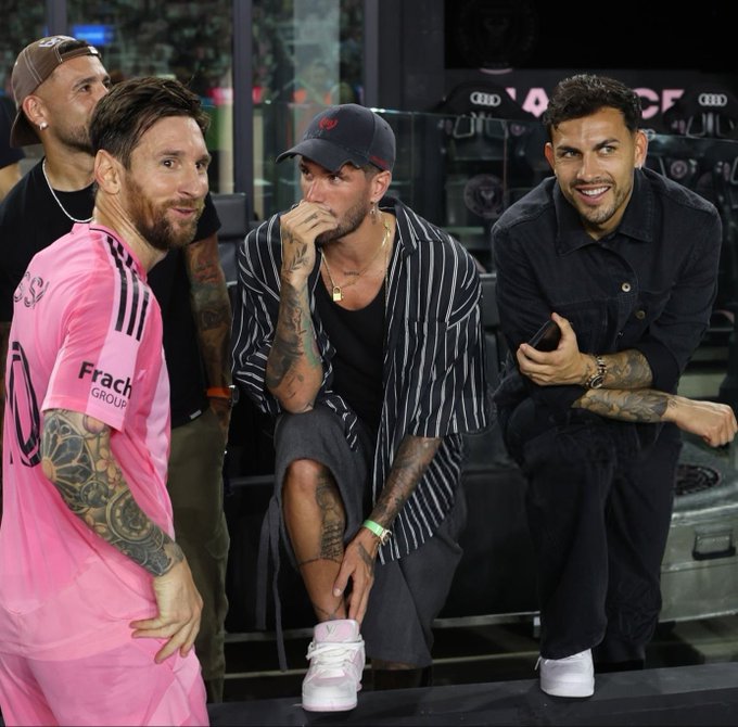 Four men stand casually on a stage-like area with a black backdrop displaying the Audi logo and sponsor branding. Lionel Messi, wearing a pink Adidas jersey with number 10 and sponsor patches, smiles while standing with arms at sides. To his left, a man in a black cap and striped shirt rubs his nose thoughtfully. To his right, Julian Alvarez in a black cap and dark shirt smiles while holding a phone, and Enzo Fernandez in a black shirt grins with hand on hip. All wear casual pants, sneakers, and visible tattoos on arms; the setting suggests a post-match or event photo.