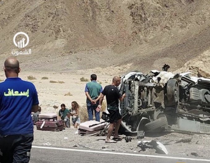 First image displays a desert road scene with a severely damaged white vehicle crashed on the roadside surrounded by scattered luggage and debris under a mountainous backdrop several people including men in casual clothing and a woman stand nearby examining the wreckage with Arabic text overlays and circular logo visible on the vehicle and ground Second image shows a nighttime view of a dark damaged car with front-end collision evident emergency lights flashing from approaching vehicles illuminating the scene Arabic text appears on the car door