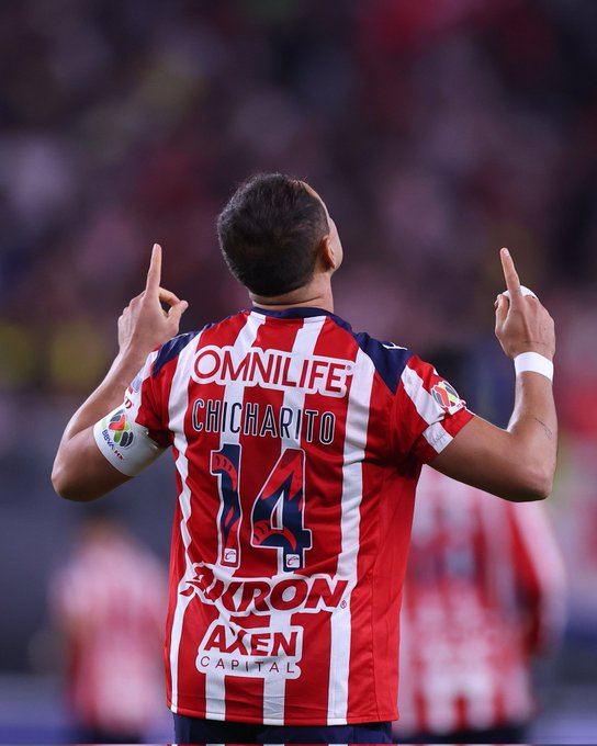 Un jugador de fútbol con cabello corto y oscuro se para en un campo en un gran estadio lleno de espectadores que visten una camiseta de Chivas a rayas rojas y blancas con el número 14 y el nombre Chicharito en la espalda patrocinado por Omnilife y Akron, levanta ambos brazos en celebración con los dedos índices apuntando hacia arriba, la camiseta tiene parches de patrocinadores como A xen Capital en la manga y el emblema del equipo en el pecho, el fondo muestra asientos borrosos del estadio y campo.