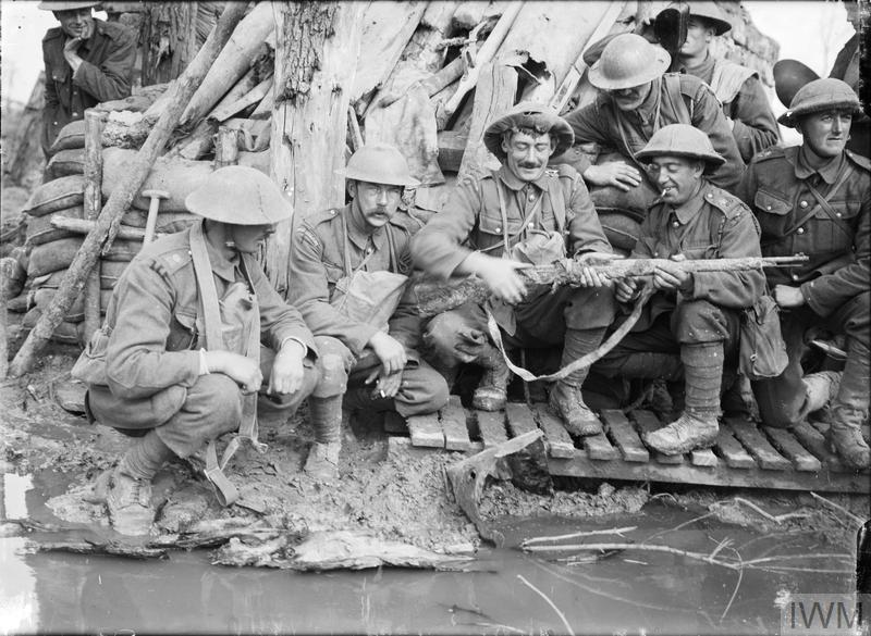A group of guards, including Coldstream Guards and Irish Guards, crouching outside a captured German dugout, examining a muddy German rifle, near Langemarck (Langemark-Poelkpelle), 12 October 1917. IWMQ3011
