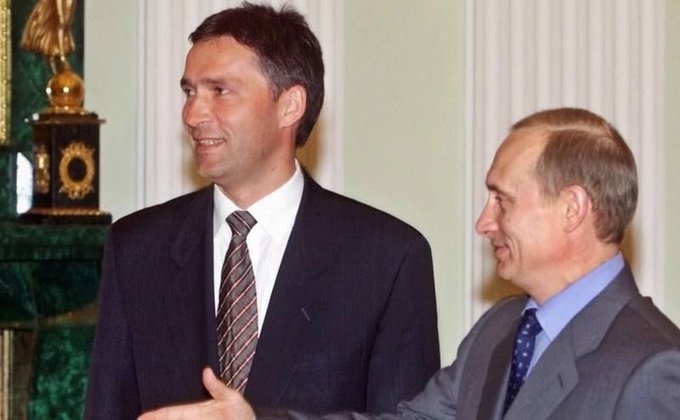 First image shows two men in suits standing side by side in an ornate room with green decor and golden lamps one with dark hair wearing a striped tie smiling slightly the other with lighter hair in a blue tie looking sideways both appearing formal and engaged in conversation. Second image depicts a group of men in suits walking in a lit hallway one central figure with glasses wearing a dark blue mask and suit others masked or unmasked beside him near a large framed portrait of an older man on the wall suggesting a formal indoor setting.
